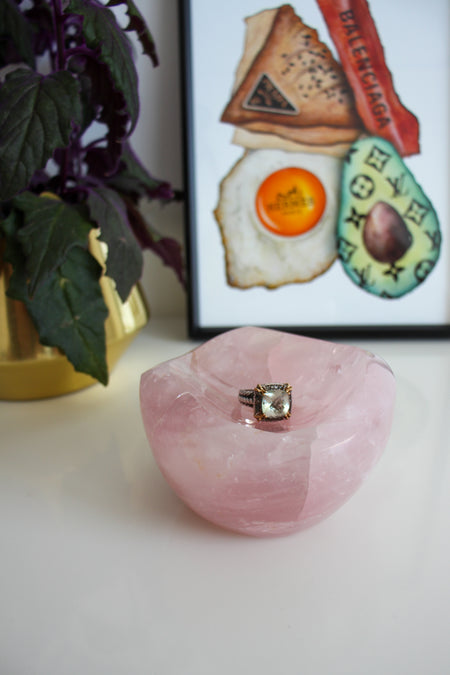A light pink rose quartz bowl sits on a white table. There is a ring with a large green stone in the bowl. Behind the bowl is a green and purple plant in a gold pot on the left. On the right behind the bowl is a framed print called 'Boujee Brunch' 