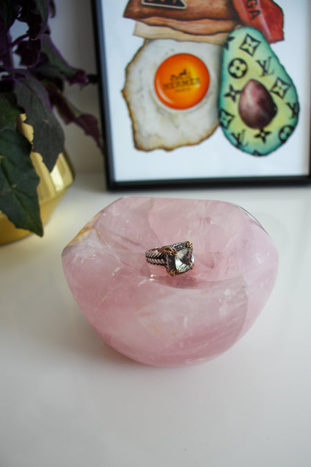 A light pink rose quartz bowl sits on a white table. There is a ring with a large green stone in the bowl. Behind the bowl is a green and purple plant in a gold pot on the left. On the right behind the bowl is a framed print called 'Boujee Brunch'