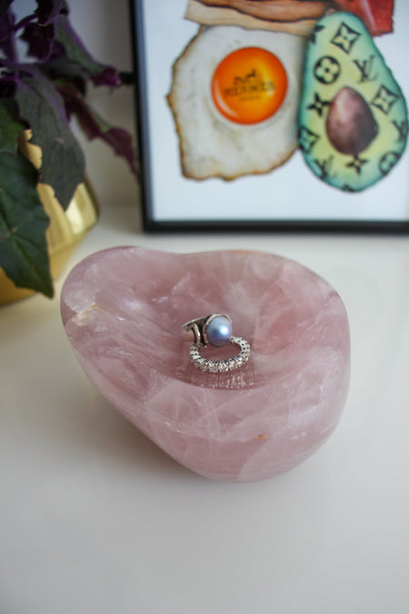 A dark pink rose quartz bowl sits on a white table. There are two rings, one a blue pearl ring and one diamond eternity ring in the bowl. Behind the bowl is a green and purple plant in a gold pot on the left. On the right behind the bowl is a framed print called 'Boujee Brunch'