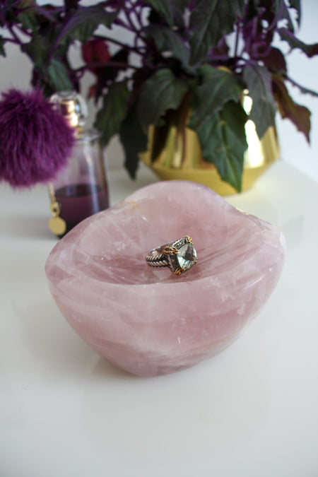 A dark pink rose quartz bowl sits on a white table. On the right behind the rose quartz bowl is a green and purple plant in a gold pot. On the left, behind the rose quartz bowl is a bottle of perfume, with a purple pom pom and it contains purple liquid. In the rose quartz bowl sits a fancy cocktail ring with a huge pale green faceted stone. 