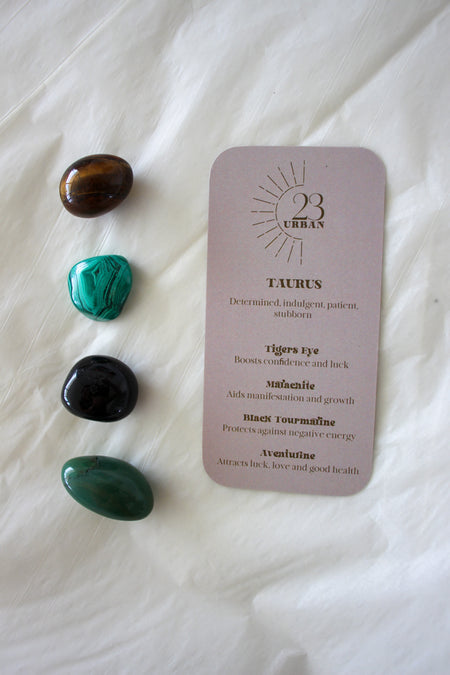 To the left of this image are the four Taurus Zodiac Crystal Kit tumble stones, which are brown and yellow Tigers Eye, green Malachite, Black Tourmaline and green Aventurine. Next to the tumble stones is a Taurus Zodiac Crystal Kit card, outlining the traits of Taurus and explaining the properties of the crystals. 