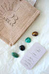 This image shows the 23 Urban Taurus Zodiac Kit in flat lay. To the upper leeft of the picture is the 23 Urban jute bag which all products come in. Then are the four Taurus tumble stones, which are brown and yellow Tigers Eye, green Malachite, Black Tourmaline and green Aventurine. Next to the tumble stones on the bottom right is the Taurus Zodiac Crystal Kit card, outlining the traits of Taurus and explaining the properties of the crystals.  