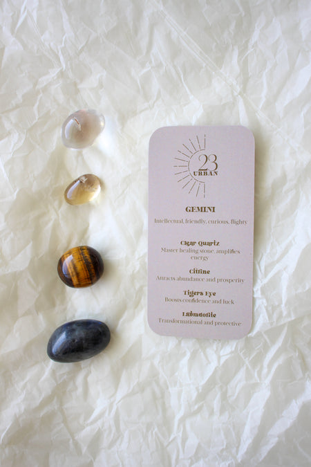 To the left of this image are the four Gemini Zodiac Crystal Kit tumble stones, which are Clear Quartz, yellow Citrine, brown and yellow Tigers Eye and green Aventurine. Next to the tumble stones is a Gemini Zodiac Crystal Kit card, outlining the traits of Gemini and explaining the properties of the crystals. 