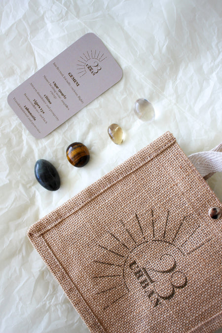  This image shows the 23 Urban Gemini Zodiac Kit in flat lay. To the bottom right of the picture is the 23 Urban jute bag which all products come in. Then are the four Gemini tumble stones, which are Clear Quartz, yellow Citrine, brown and yellow Tigers Eye and Labradorite. Next to the tumble stones on the top left is the Gemini Zodiac Crystal Kit card, outlining the traits of Gemini and explaining the properties of the crystals. 