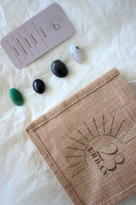  This image shows the 23 Urban Cancer Zodiac Kit in flat lay. On the bottom right is the 23 Urban jute bag which all products come in. Then are the four Cancer tumble stones, which are Moonstone, Labradorite, Black Tourmaline and green Aventurine. Next to the tumble stones on the bottom right is the Cancer Zodiac Crystal Kit card, outlining the traits of Cancer and explaining the properties of the crystals. 