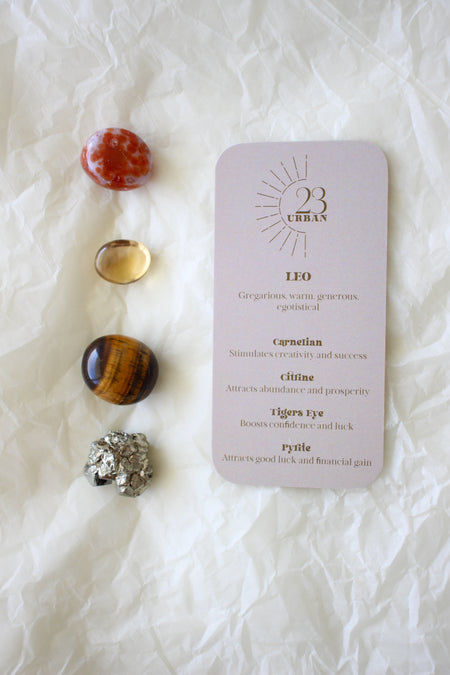 To the left of this image are the four Leo Zodiac Crystal Kit tumble stones, which are Carnelian, Citrine, Tigers Eye, Pyrite. Next to the tumble stones is a Leo Zodiac Crystal Kit card, outlining the traits of Leo and explaining the properties of the crystals. 