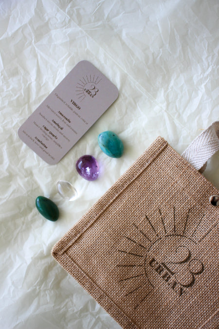 This image shows the 23 Urban Virgo Zodiac Kit in flat lay. On the bottom right is the 23 Urban jute bag which all products come in. Then are the four Virgo tumble stones, which are Amazonite, Amethyst, Clear Quartz and Aventurine. Next to the tumble stones on the bottom right is the Virgo  Zodiac Crystal Kit card, outlining the traits of Virgo and explaining the properties of the crystals. 