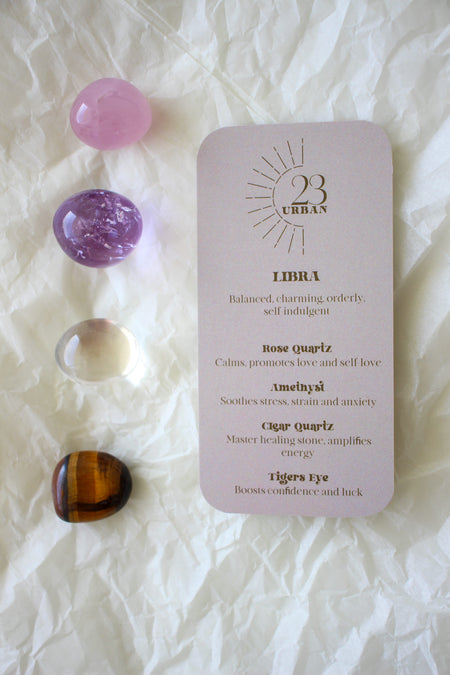 To the left of this image are the four Libra Zodiac Crystal Kit tumble stones, which are Rose Quartz, Amethyst, Clear Quartz and Tigers Eye. Next to the tumble stones is a Libra Zodiac Crystal Kit card, outlining the traits of Libra and explaining the properties of the crystals. 