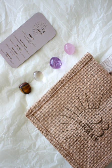 This image shows the 23 Urban Libra Zodiac Kit in flat lay. On the bottom right is the 23 Urban jute bag which all products come in. Then are the four Libra tumble stones, which are Rose Quartz, Amethyst, Clear Quartz and Tigers Eye. Next to the tumble stones on the bottom right is the Libra Zodiac Crystal Kit card, outlining the traits of Libra and explaining the properties of the crystals. 