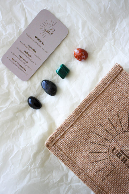 This image shows the 23 Urban Scorpio Zodiac Kit in flat lay. On the bottom right is the 23 Urban jute bag which all products come in. Then are the four Scorpio tumble stones, which are Carnelian, Malachite, Labradorite and Black Tourmaline. Next to the tumble stones on the bottom right is the Scorpio Zodiac Crystal Kit card, outlining the traits of Libra and explaining the properties of the crystals.