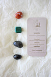 To the left of this image are the four Scorpio Zodiac Crystal Kit tumble stones, which are Carnelian, Malachite, Labradorite and Black Tourmaline. Next to the tumble stones is a Scorpio Zodiac Crystal Kit card, outlining the traits of Scorpio and explaining the properties of the crystals. 