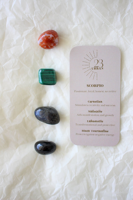 To the left of this image are the four Scorpio Zodiac Crystal Kit tumble stones, which are Carnelian, Malachite, Labradorite and Black Tourmaline. Next to the tumble stones is a Scorpio Zodiac Crystal Kit card, outlining the traits of Scorpio and explaining the properties of the crystals. 
