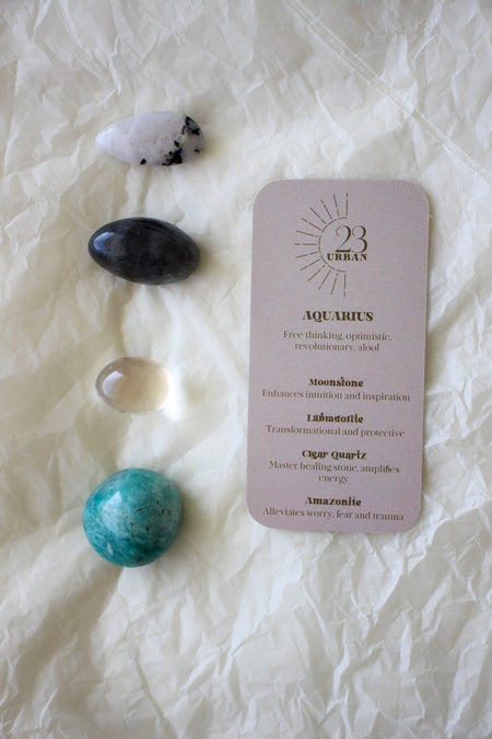 To the left of this image are the four Aquarius Zodiac Crystal Kit tumble stones, which are Moonstone, Labradorite, Clear Quartz, Amazonite. Next to the tumble stones is an Aquarius Zodiac Crystal Kit card, outlining the traits of Aquarius and explaining the properties of the crystals. 