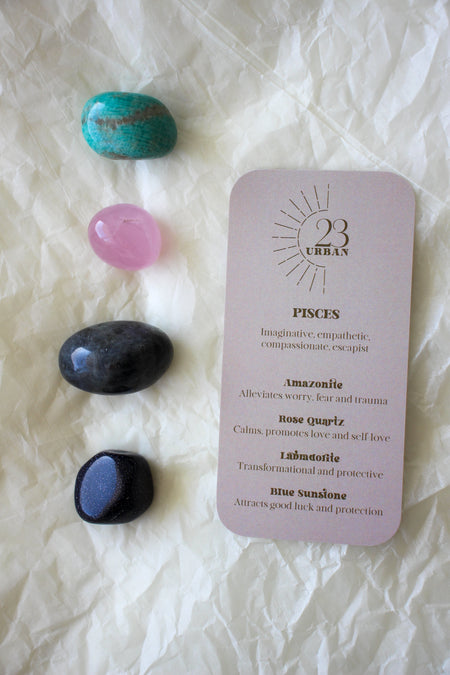 To the left of this image are the four Pisces Zodiac Crystal Kit tumble stones, which are Amazonite, Rose Quartz, Labradorite, Blue Sunstone. Next to the tumble stones is a Pisces Zodiac Crystal Kit card, outlining the traits of Pisces and explaining the properties of the crystals. 