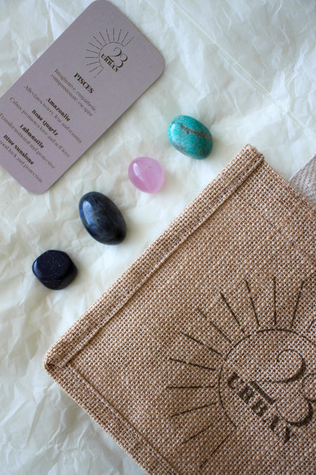  This image shows the 23 Urban Pisces Zodiac Kit in flat lay. On the bottom right is the 23 Urban jute bag which all products come in. Then are the four Pisces tumble stones, which are Amazonite, Rose Quartz, Labradorite, Blue Sunstone. Next to the tumble stones on the bottom right is the Pisces Zodiac Crystal Kit card, outlining the traits of Pisces and explaining the properties of the crystals. 