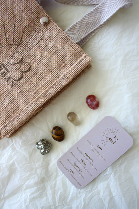 This image shows the 23 Urban Leo Zodiac Kit in flat lay. On the top left is the 23 Urban jute bag which all products come in. Then are the four Leo tumble stones, which are Carnelian, Citrine, Tigers Eye, Pyrite. Next to the tumble stones on the bottom right is the Leo Zodiac Crystal Kit card, outlining the traits of Leo and explaining the properties of the crystals. 