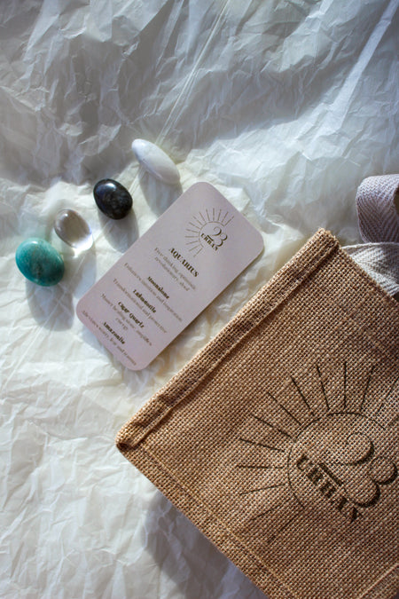  This image shows the 23 Urban Aquarius Zodiac Kit in flat lay. On the top left are the four Aquarius tumble stones, which are Moonstone, Labradorite, Clear Quartz, Amazonite.   Next to the tumblestones is the Aquarius Zodiac Crystal Kit card, outlining the traits of Aquarius and explaining the properties of the crystals. Then on the bottom right is the 23 Urban jute bag which all products come in.