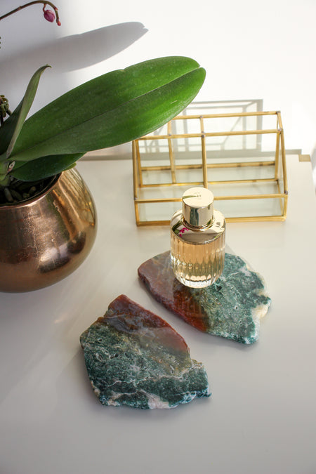 Two green and red Ocean Jasper slabs sit on a white table. On one slab is a bottle of perfume. In the background is a makeup brush holder and to the left is an orchid in a bronze pot. 