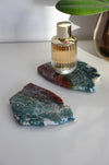 Two red and green Ocean Jasper slabs sit on a white table. On one slab is a bottle of perfume with a gold lid. 