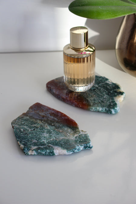 Two red and green Ocean Jasper slabs sit on a white table. On one slab is a bottle of perfume with a gold lid. 