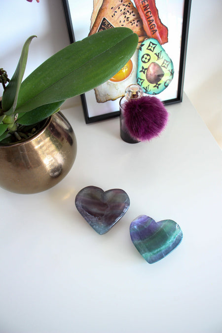 Two Rainbow Fluorite bowls sit on a white desk in front of a bottle of perfume, an orchid plant and a painting. The two bowls are heart shaped and semi transparent. Their colours are purple, green and blue 