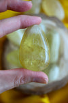 A pale lemon yellow citrine palm stone is held between two fingers, in the background is a yellow cloth 