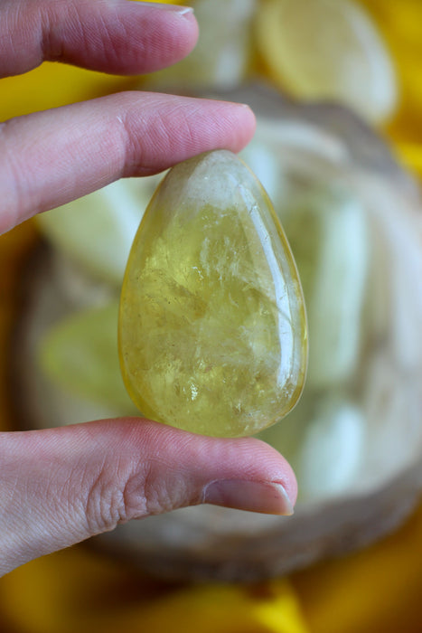 A pale lemon yellow citrine palm stone is held between two fingers, in the background is a yellow cloth 