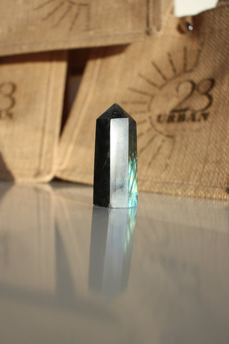 A Labradorite Tower crystal with an intense blue flash stands on a white table. In the background are 23 Urban branded jute bags. 