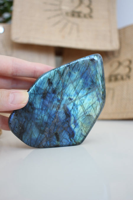 A Labradorite Freeform crystal with an intense blue flash stands on a white table. In the background are 23 Urban branded jute bags. 