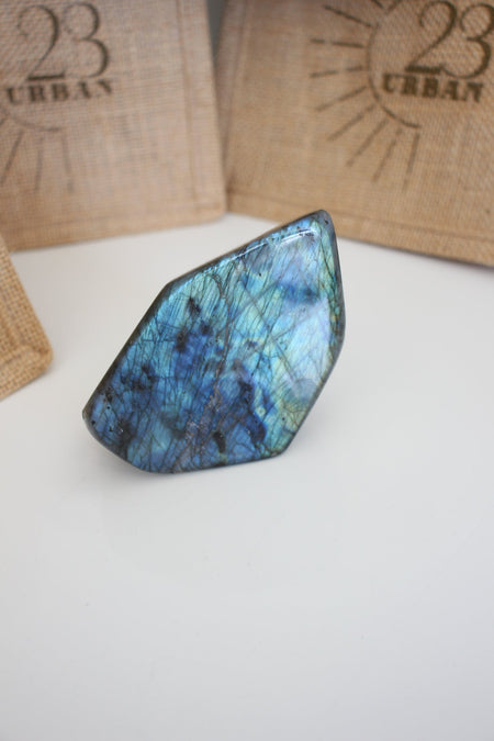 A Labradorite Freeform crystal with an intense blue flash stands on a white table. In the background are 23 Urban branded jute bags. 