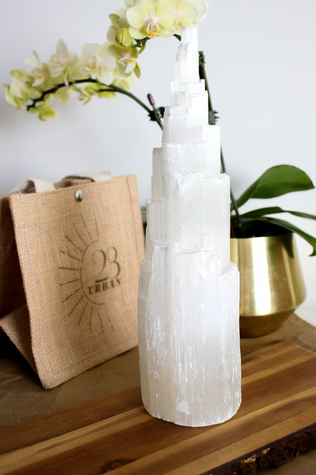 An extra large Selenite Tower stands on a brown table with a 23 Urban jute bag in the background. Also in the background is a yellow orchid in a gold pot. 