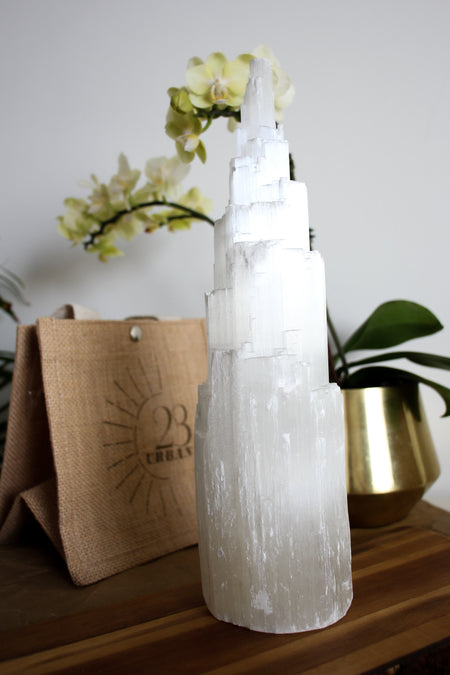 An extra large Selenite Tower stands on a brown table with a 23 Urban jute bag in the background. Also in the background is a yellow orchid in a gold pot. 