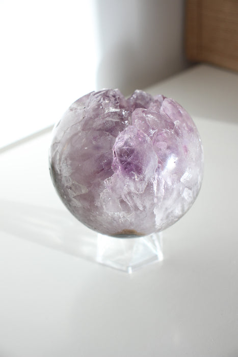 A beautiful Uruguayan Amethyst Sphere sits on a white bookshelf in the afternoon sun 