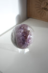 A juicy purple and white amethyst sphere sits on a white bookshelf in the afternoon sunlight