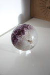 A juicy purple and white amethyst sphere sits on a white bookshelf in the afternoon sunlight