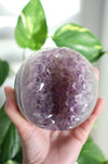 A large juicy purple Amethyst Sphere is held up by a hand in the afternoon sunlight