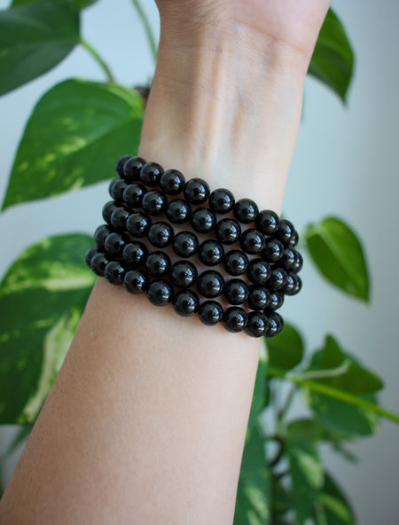 A stack of shiny Black Tourmaline bracelets on a wrist in front of a green plant 