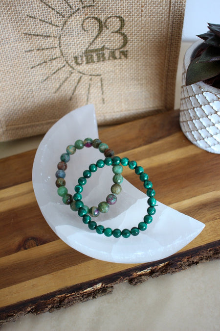 A white selenite moon shaped bowl sits on a brown table. Inside it is a malachite bracelet and a ruby-in-zoisite bracelet