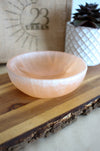 A peach selenite cleansing bowl sits on a brown table with a plant in the background 