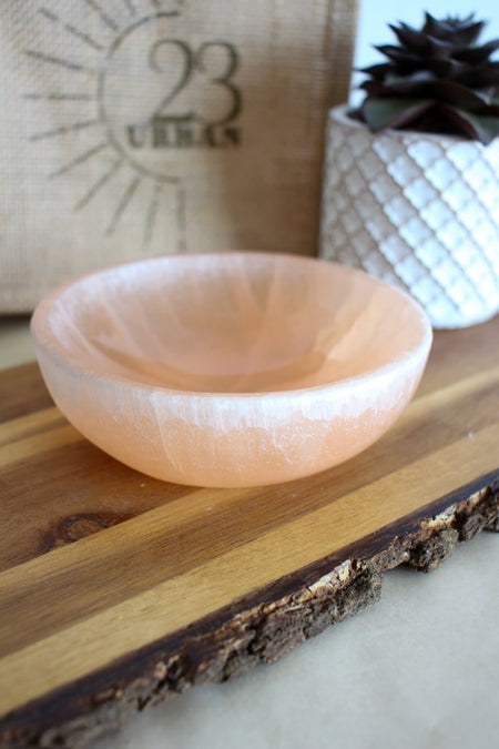 A peach selenite cleansing bowl sits on a brown table with a plant in the background 