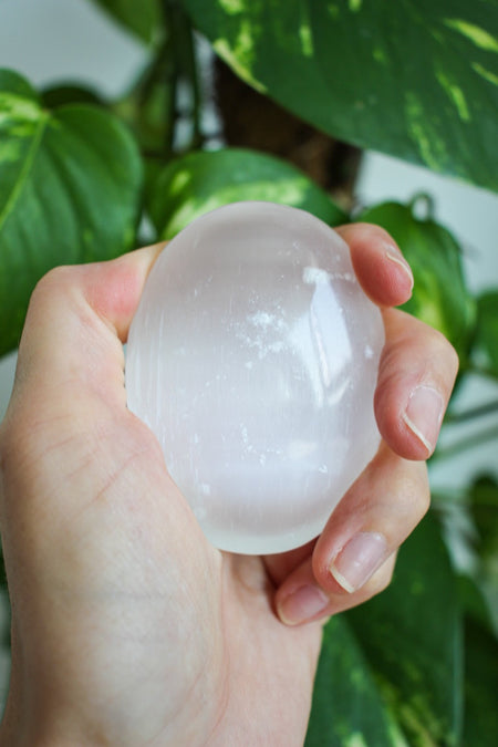 A glowing white selenite palm stone sits in a persons hand. in the background is a green plant