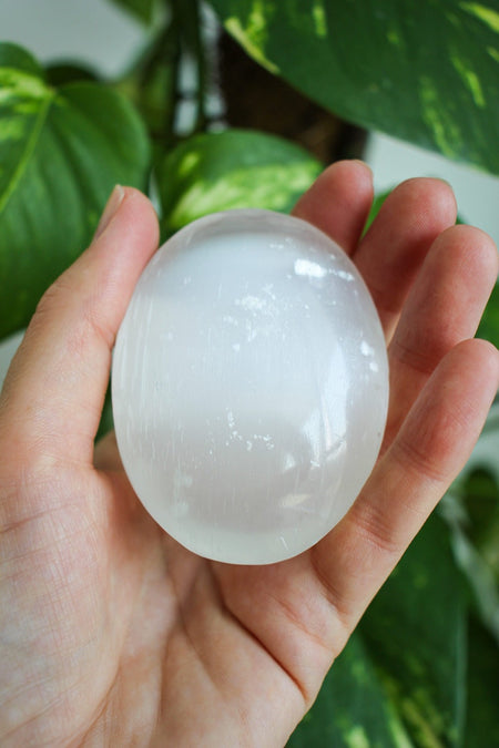 A glowing white selenite palm stone sits in a persons hand. in the background is a green plant 