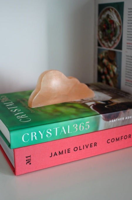 A peach coloured, cloud shaped selenite cleansing plate sits upright on a pile of books