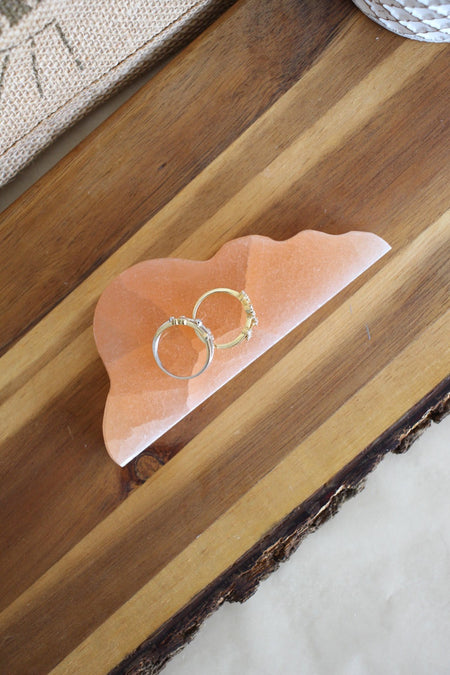 A peach coloured, cloud shaped cleansing plate sits flat on a brown table. On it sits two gold rings