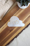 A cloud shaped white selenite cleansing plate sits on a brown table, it has two gold rings on it 