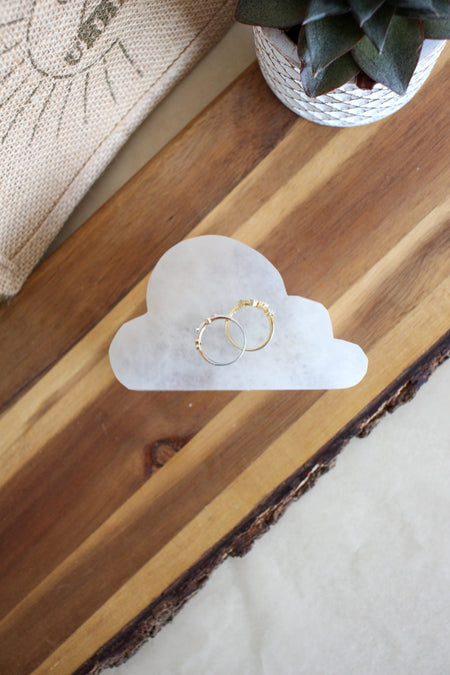 A cloud shaped white selenite cleansing plate sits on a brown table, it has two gold rings on it 