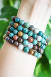 Beautiful blue and brown toned Ocean Jasper, pictured stacked on a wrist in front of a green plant