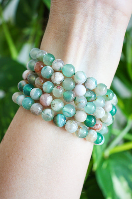 A stack of Green Flower Agate bracelets on a wrist in front of a green plant 