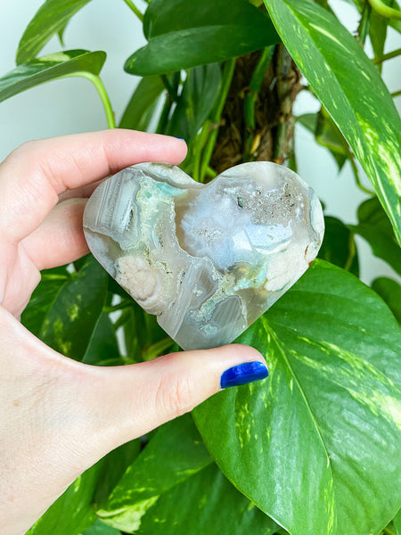 A beautiful Green Flower Agate Heart is held in front of a green plant