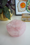 A light pink rose quartz bowl sits on a white table. Behind the bowl is a green and purple plant in a gold pot on the left. On the right behind the bowl is a framed print called 'Boujee Brunch'